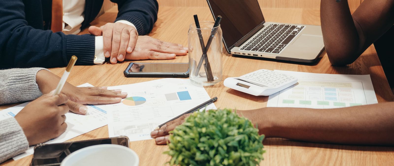 Three colleagues around a meeting table reviewing printed budget reports and charts, with a laptop, calculator and notebooks open.